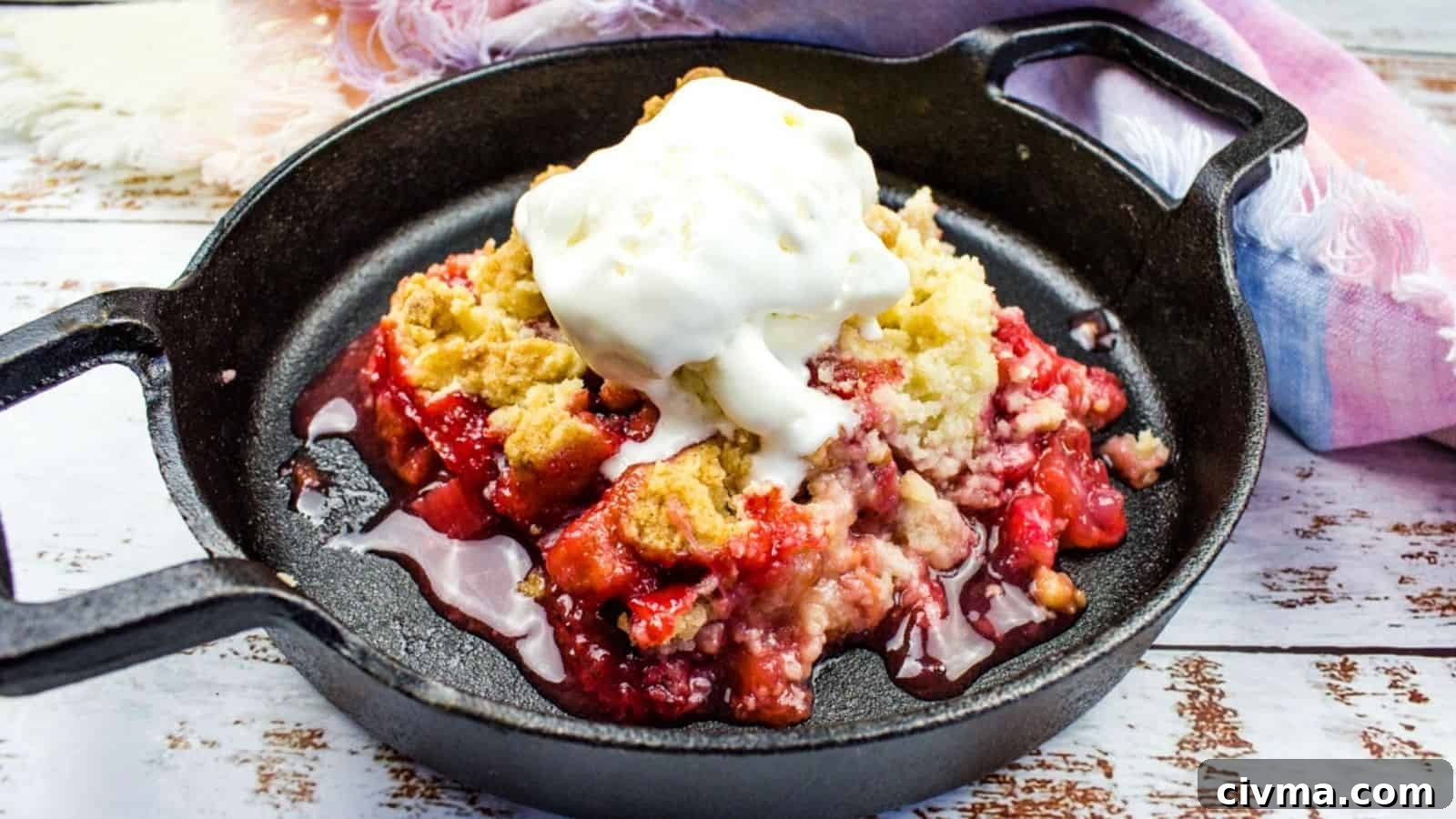 A cast iron skillet with strawberry rhubarb cobbler topped with a scoop of vanilla ice cream on a wooden surface with a cloth napkin beside it.