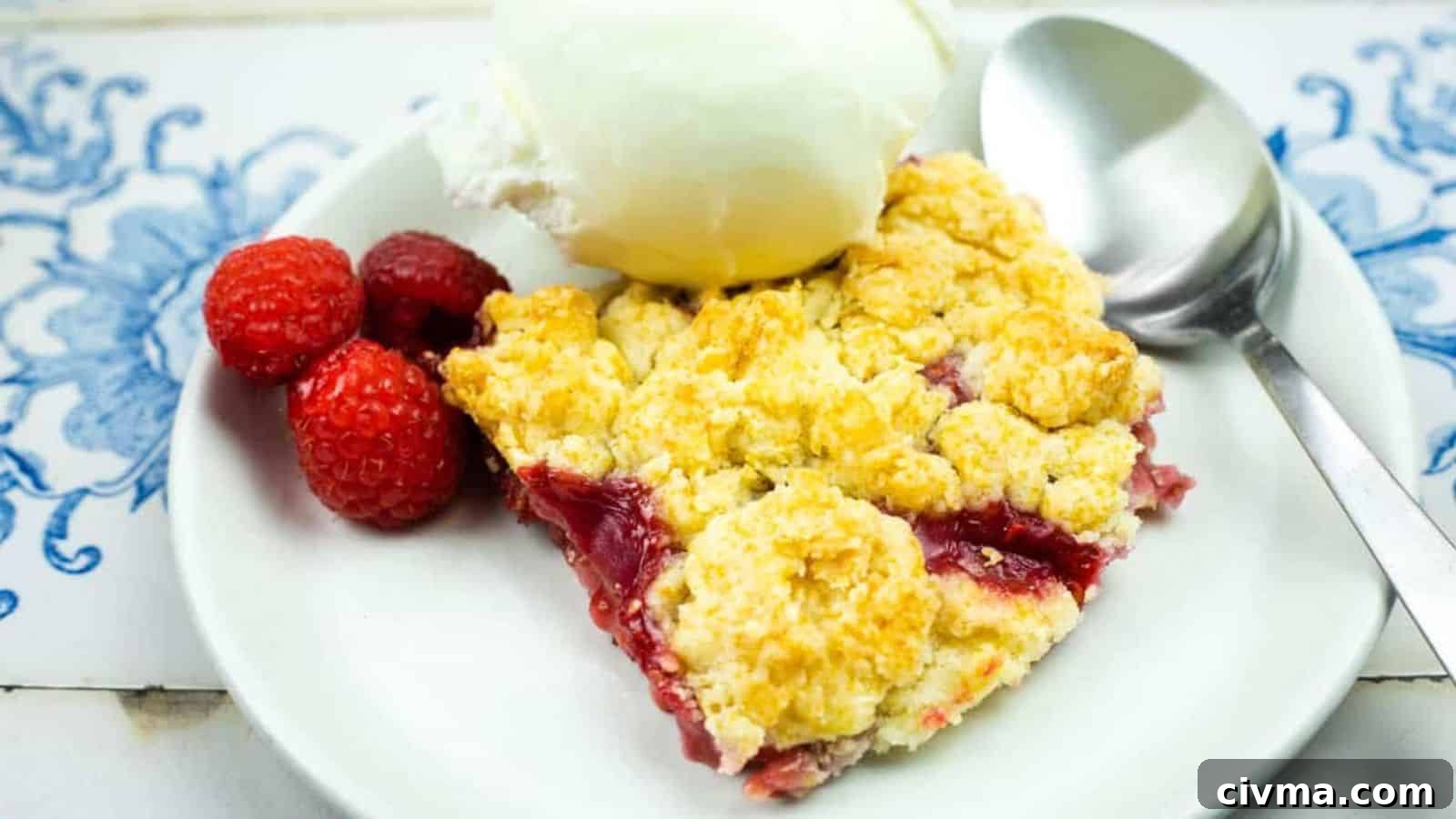 A slice of berry cobbler topped with a scoop of vanilla ice cream is served on a white plate with a spoon and fresh raspberries.