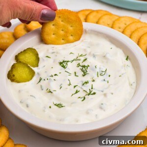 Small white bowl filled with creamy white dip, topped with slices of pickles, surrounded by round crackers, and a plate of fresh vegetables.