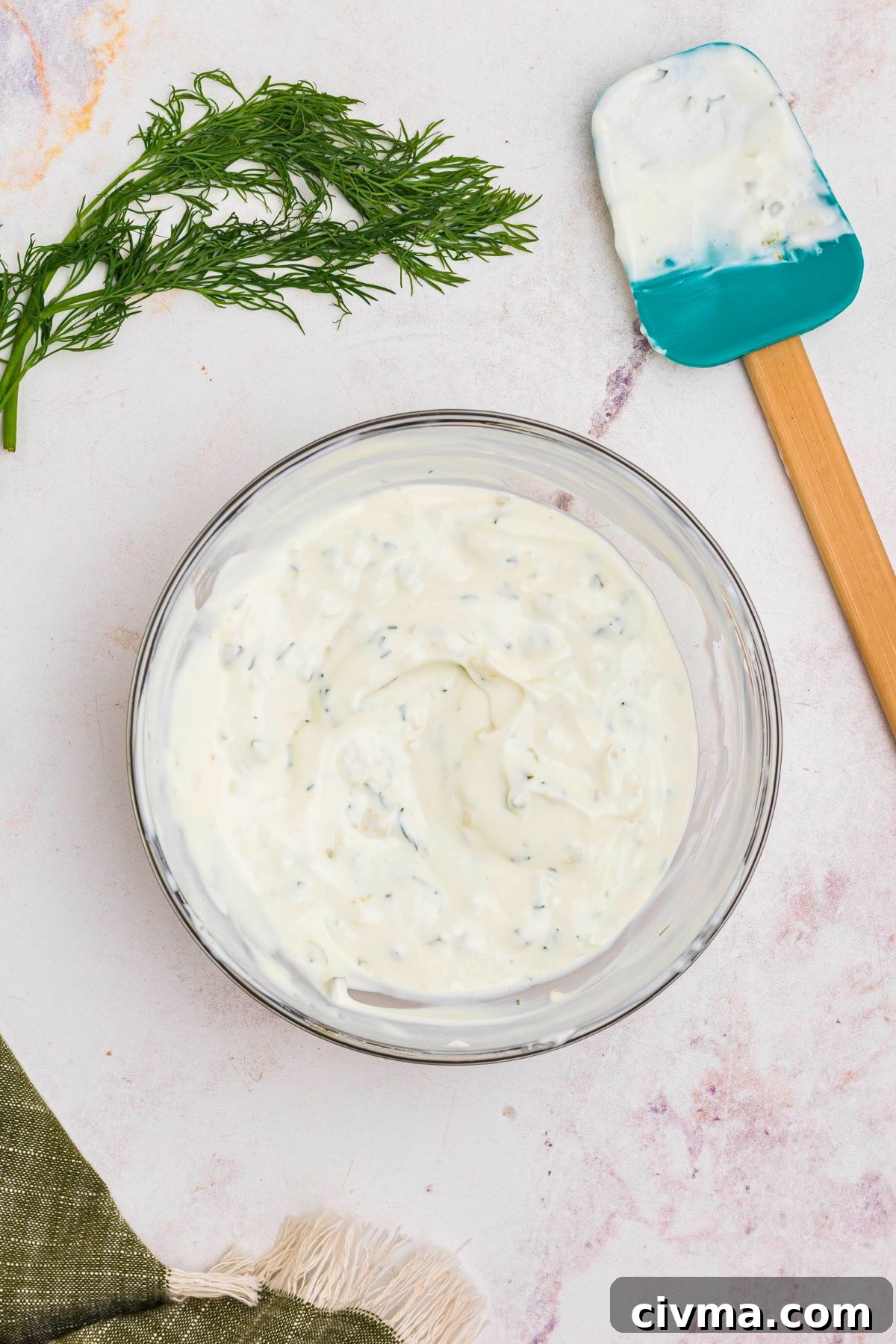 Creamy dip mixed together in a clear glass bowl with a spatula and fresh dill laying on the table next to the bowl. 