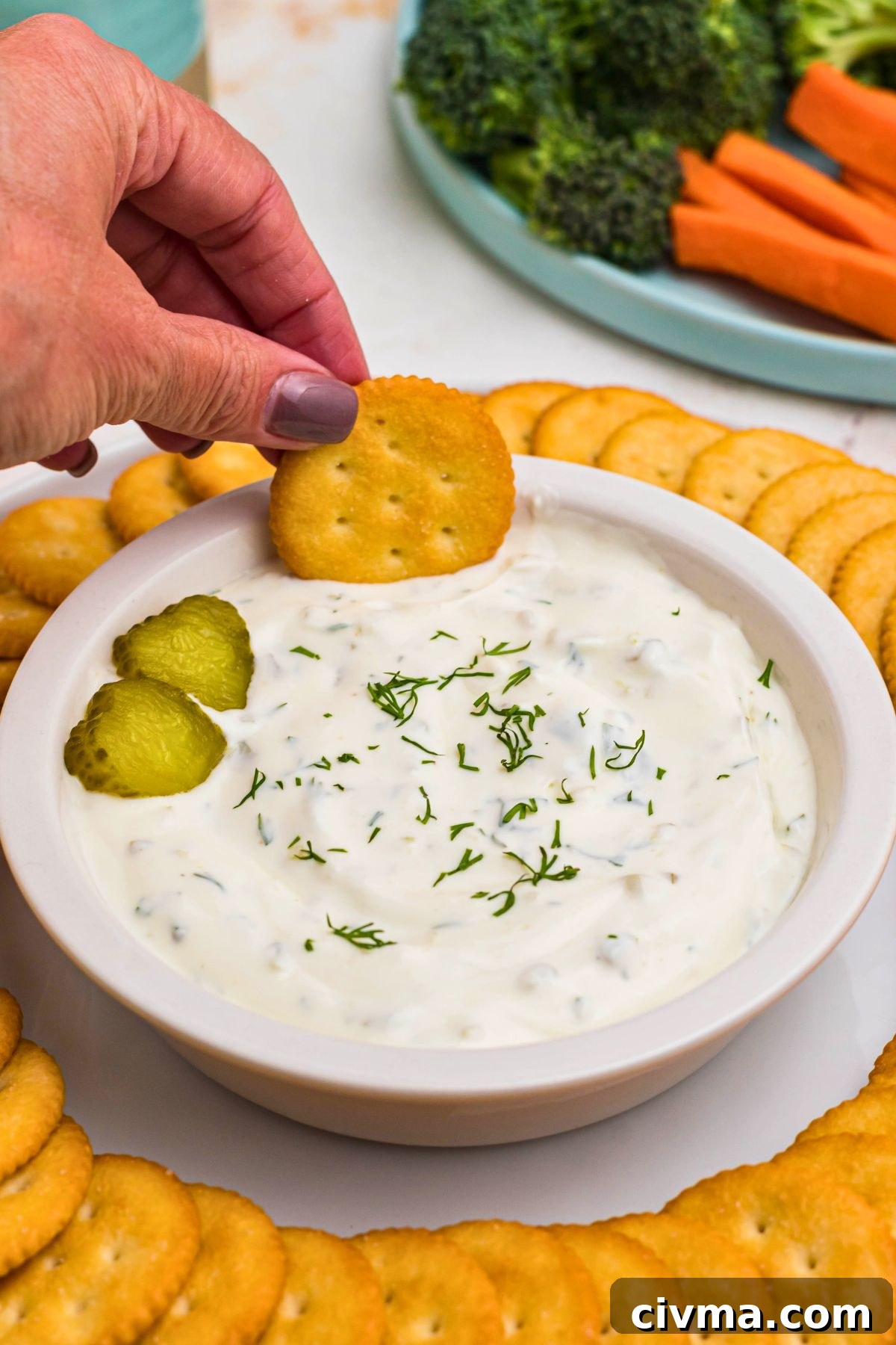 Small white bowl filled with creamy white dip, topped with slices of pickles, surrounded by round crackers, and a plate of fresh vegetables.