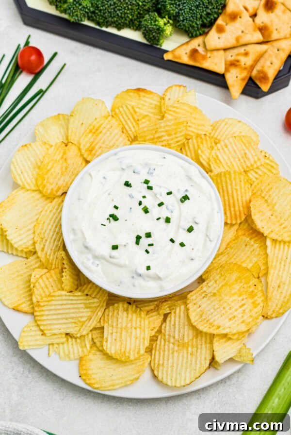 Large plate of potato chips with small round bowl in the center of chips, filled with white creamy dip, surrounded by fresh vegetables on a platter.