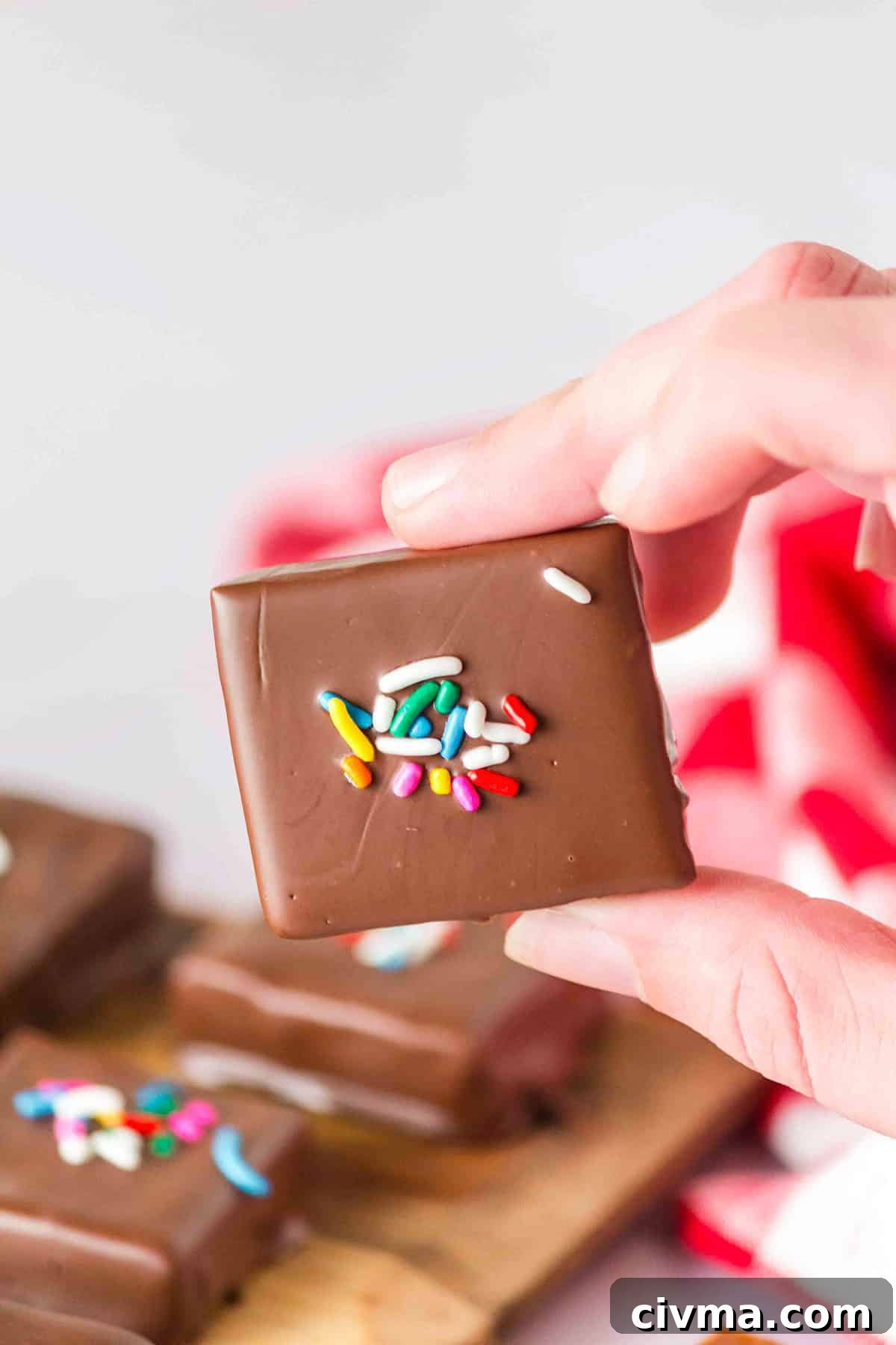 A hand holds a square, chocolate-covered Soft & Chewy Caramel treat topped with colorful sprinkles; more similar delights are visible in the background.