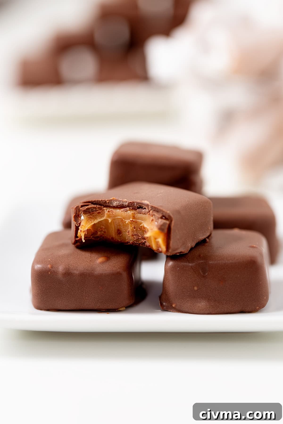 A close-up of Soft & Chewy Caramels coated in chocolate on a white plate, with one candy showing a bite that reveals the gooey caramel filling inside.