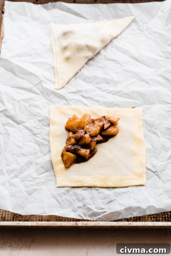 A square of puff pastry, with egg wash brushed over the edges, and apple filling on one half. A folded apple turnover is in the background on the same baking sheet