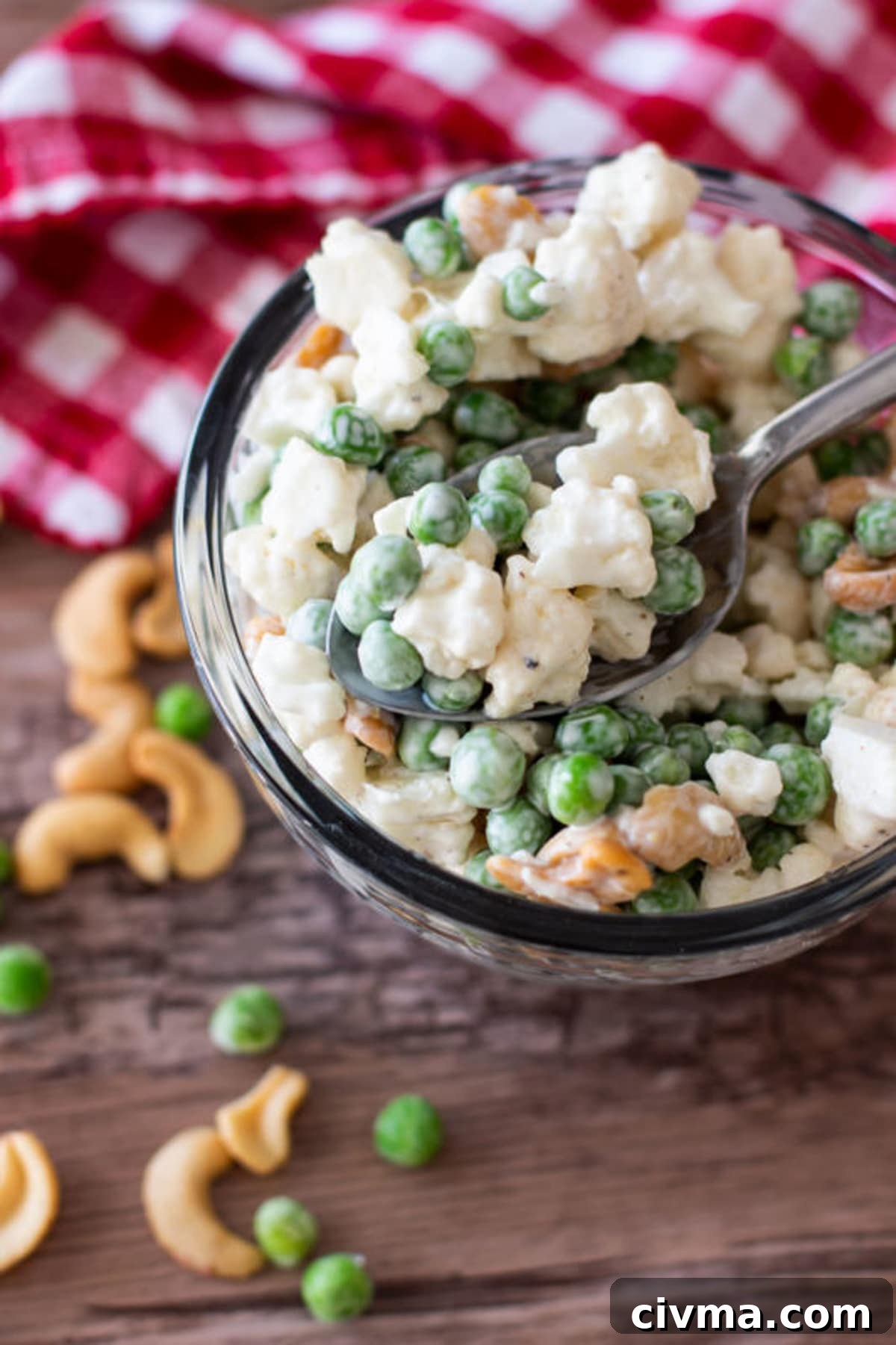 Spoonful of cauliflower and pea salad with cashews in a glass bowl sitting on a white gingham napkin, scattered peas and cashew nuts, silver spoon in bowl.