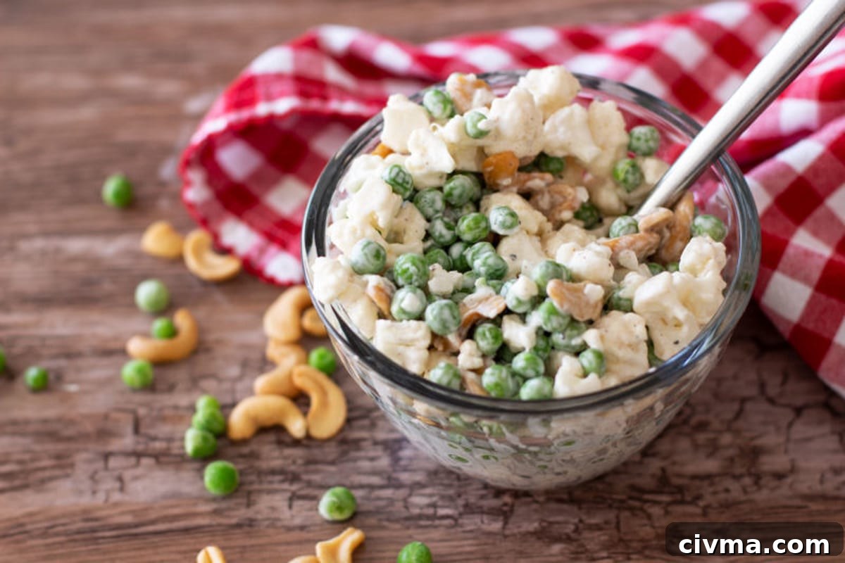 Glass bowl containing a cold pea and cauliflower salad, cashews on table.