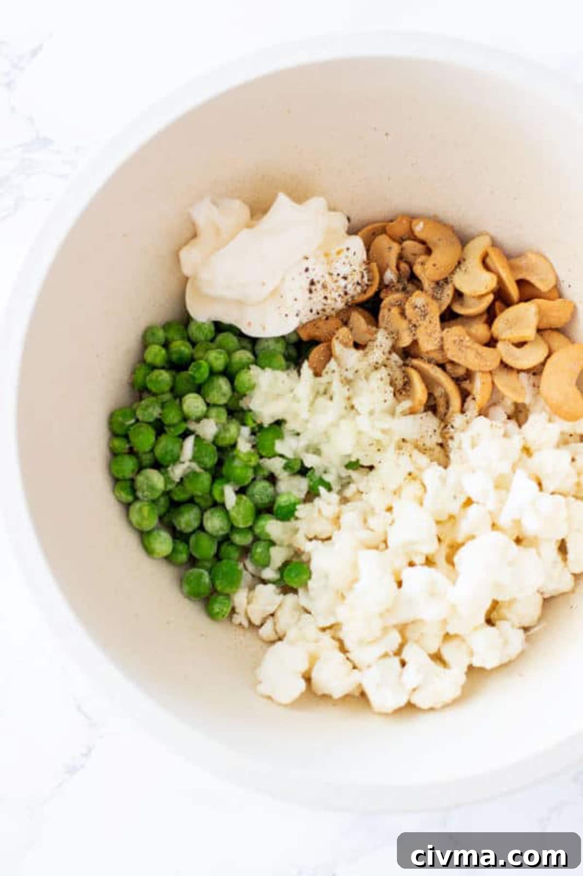 Bowl containing frozen peas, cashew halves, cauliflower, onion and mayonnaise topped with salt and pepper.