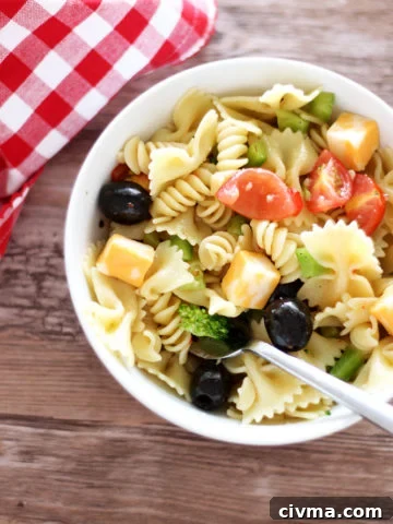White bowl containing pasta salad topped with tomatoes, peppers, cheese and broccoli, fork in bowl with red gingham napkin on brown table.