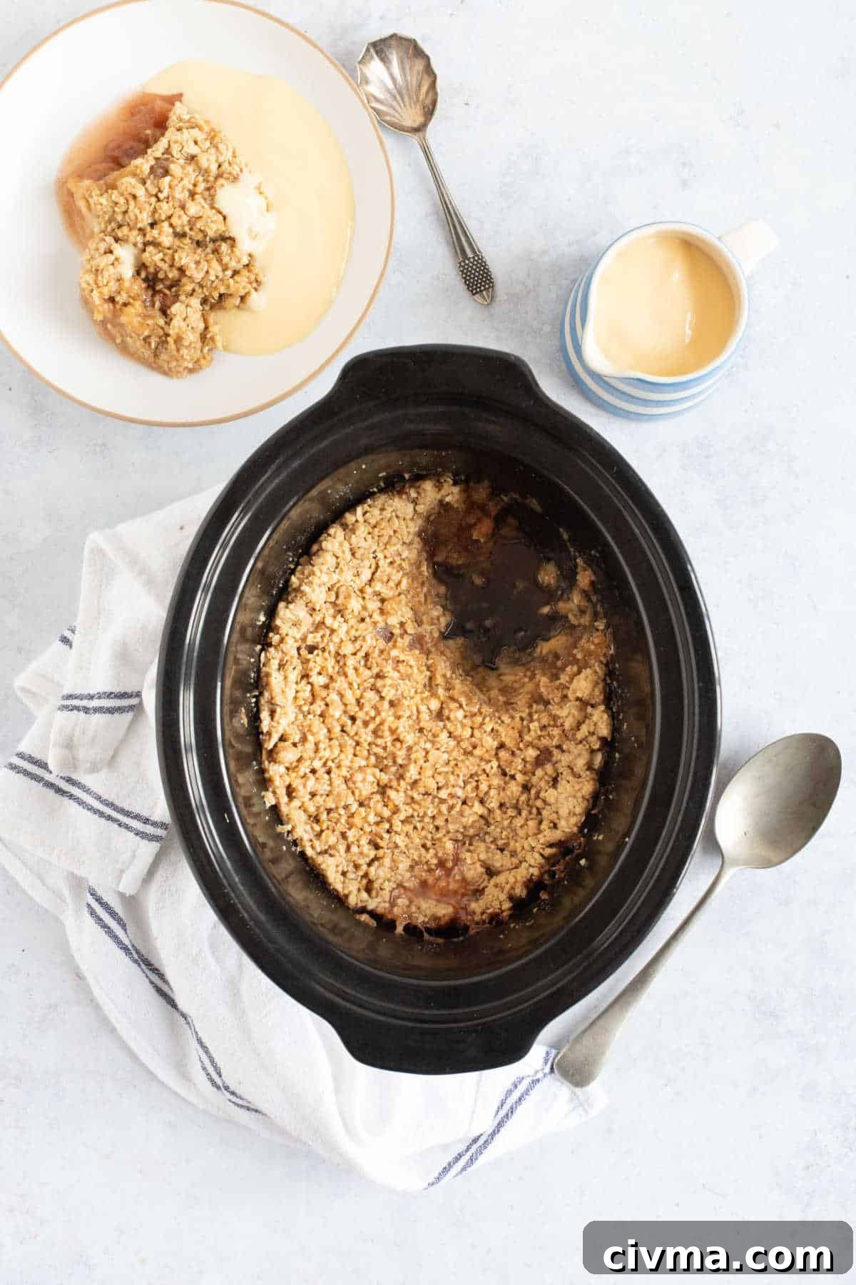 Slow cooker apple crumble in a black slow cooker basin with a bowl of apple crumble and custard on the side.