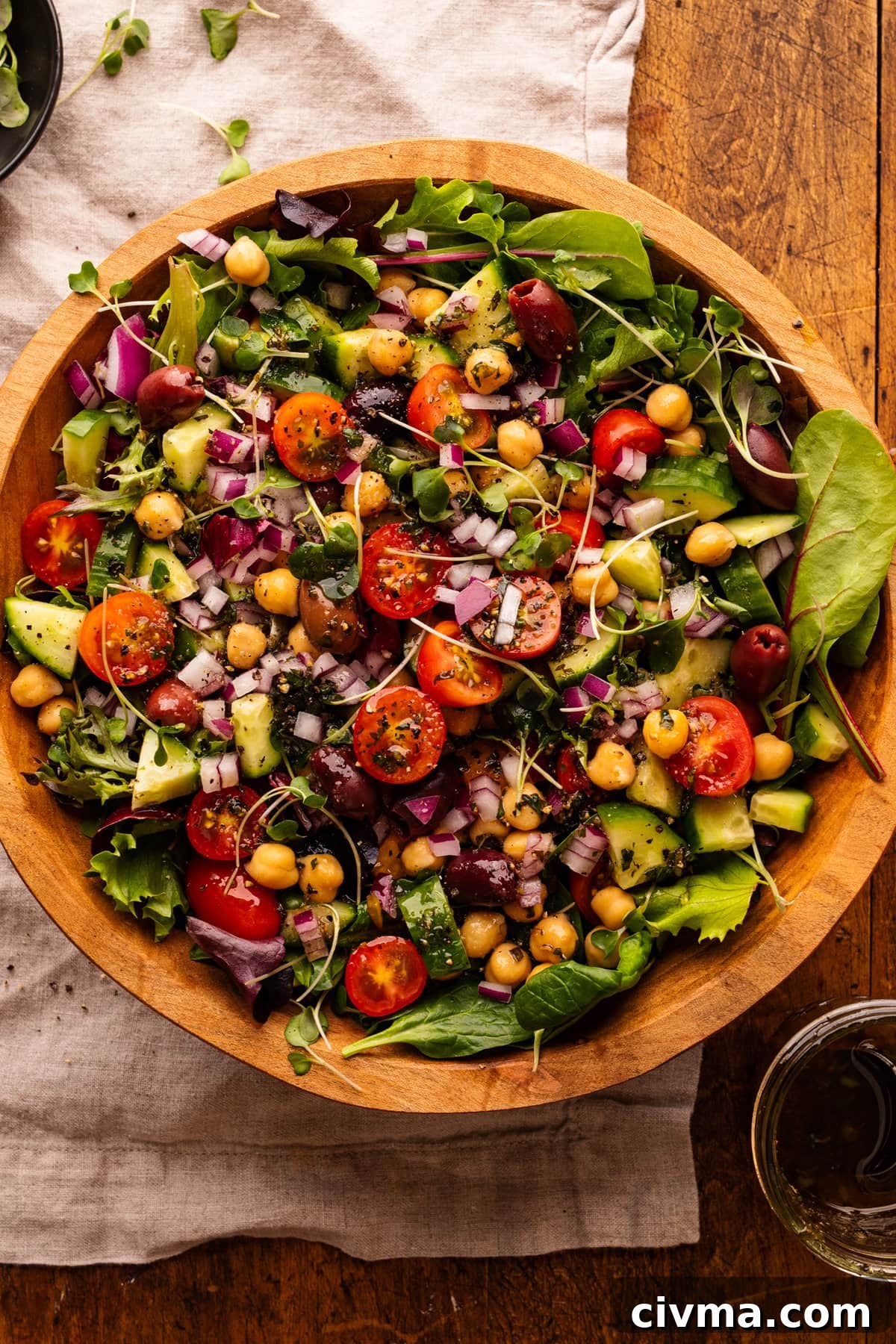 A wooden bowl with greek chickpea salad in it. The bowl is sitting on a grey napkin and wooden board.