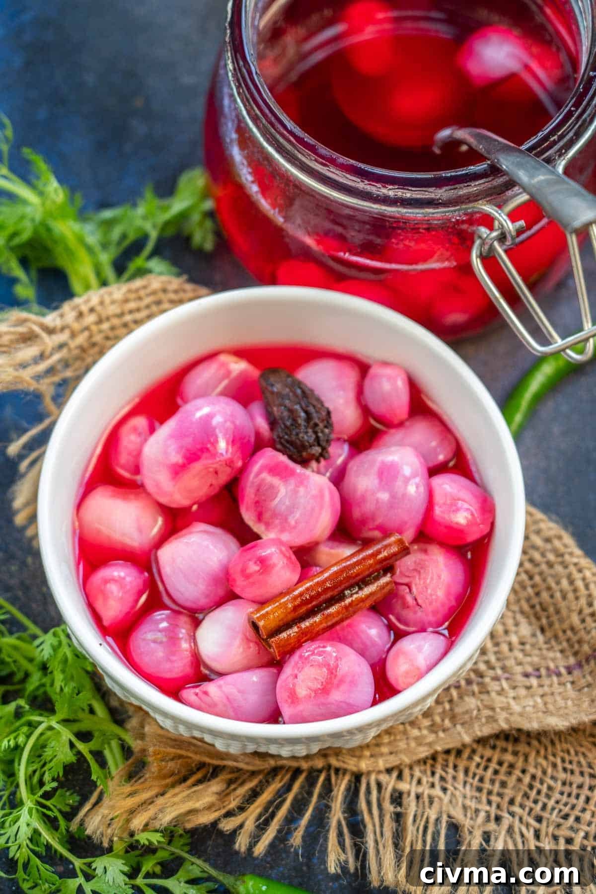 Restaurant-Style Indian Vinegar Pickled Onions 2 A white bowl filled with bright pink Sirke wale Pyaaz, a cinnamon stick, and a dark spice sits on burlap. In the background, a jar of pickling liquid and fresh herbs add to the rustic appeal.