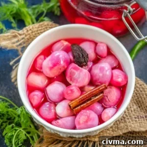 A white bowl filled with bright pink Sirke wale Pyaaz, a cinnamon stick, and a dark spice sits on burlap. In the background, a jar of pickling liquid and fresh herbs add to the rustic appeal.
