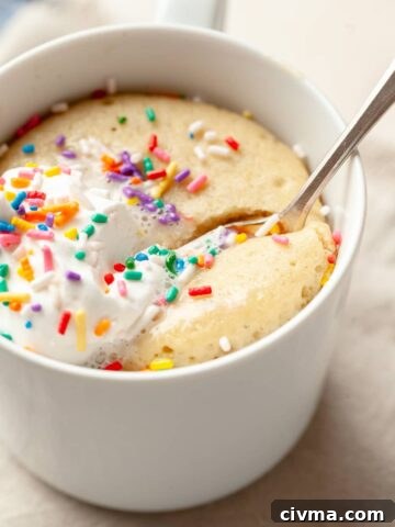 a gluten free vanilla mug cake topped with whipped cream and sprinkles in a mug, with a spoon sticking out of it.