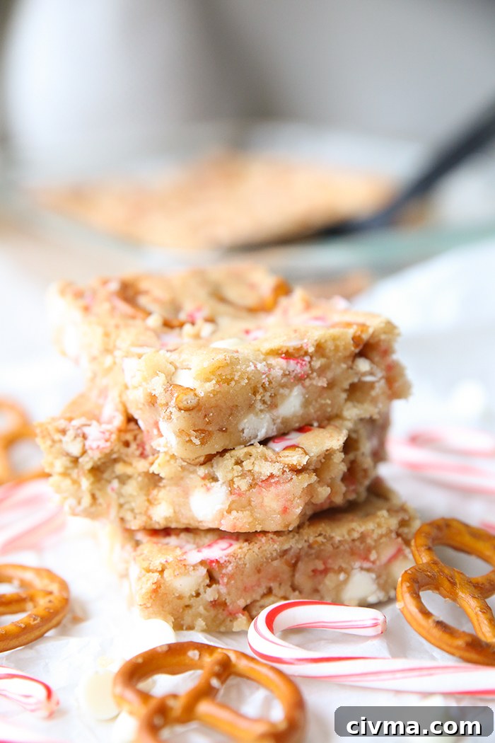 Three peppermint white chocolate pretzel cookie bars stacked on each other with small candy canes and pretzel twists surrounding them. Faded in the background is a glass 9x13 baking dish of the cookie bars and a black serving spatula.