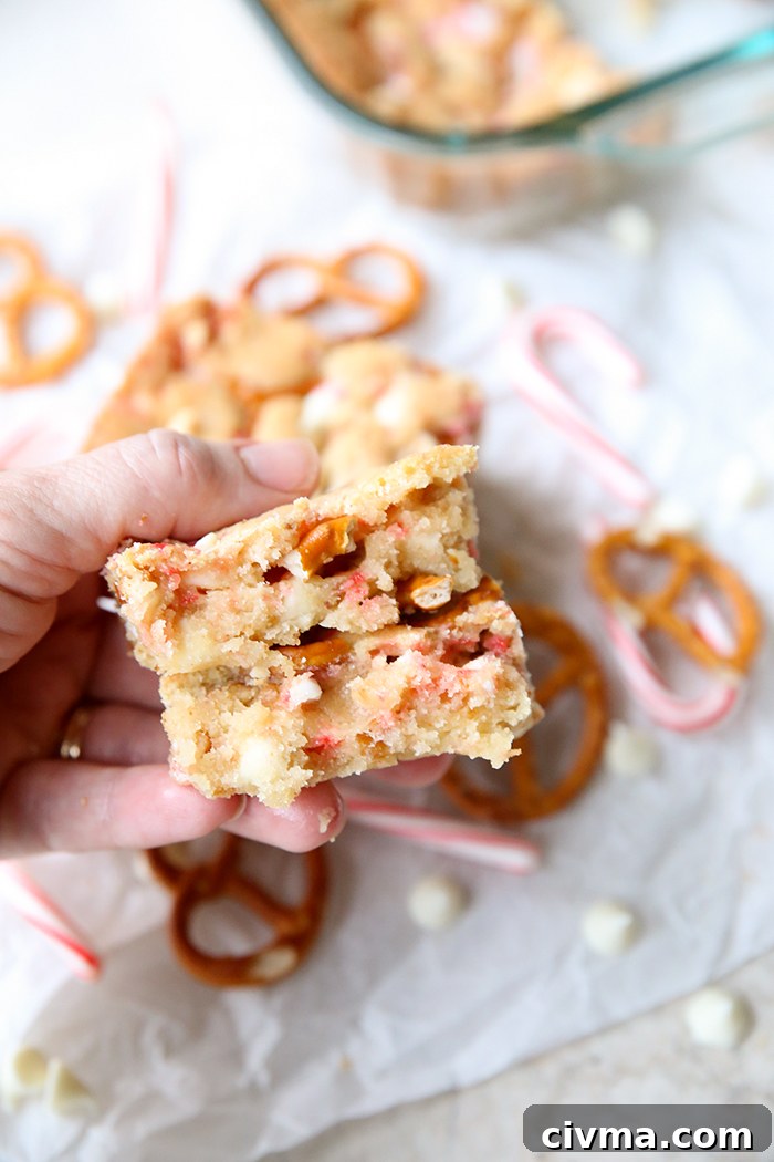 White Chocolate Peppermint Pretzel Cookie Bars 6 A broken in half white chocolate candy cane pretzel cookie bar held above scattered white chocolate chips, pretzel twists and small candy canes. In the upper right corner is a glass 9x13 dish corner with the cookie bar.