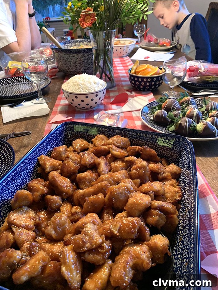 A rectangle wooden table with a red and white checkered runner down the middle with paper hearts scattered on it with a glass vase of flowers in the middle. On top of the runner is a 9x13 Polish pottery dish full of orange chicken. A plate of chocolate strawberries are placed by the right upper corner of the 9x13 dish. A white bowl with blue polka dots filled with rice is diagonally across the chocolate covered strawberries. Diagonally across from the rice bowl is a dark blue bowl with white polka dots filled with orange slices. To the left of the rice bowl is a dinner plate, a wine glass, and napkin. Diagonally to the left of the vase of flowers is a large bowl of fried rice with a wooden spoon inserted from the left. At the end of the table on the left shows a person's arms bent and the elbows resting on the table. Across from that person is a young boy looking down at a dinner plate with treats on it.