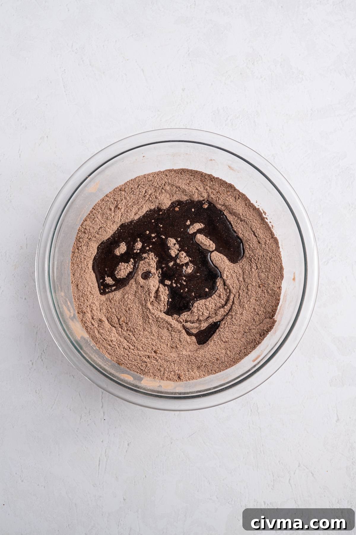 A glass bowl with cocoa powder mixture and liquid ingredients on a white surface.