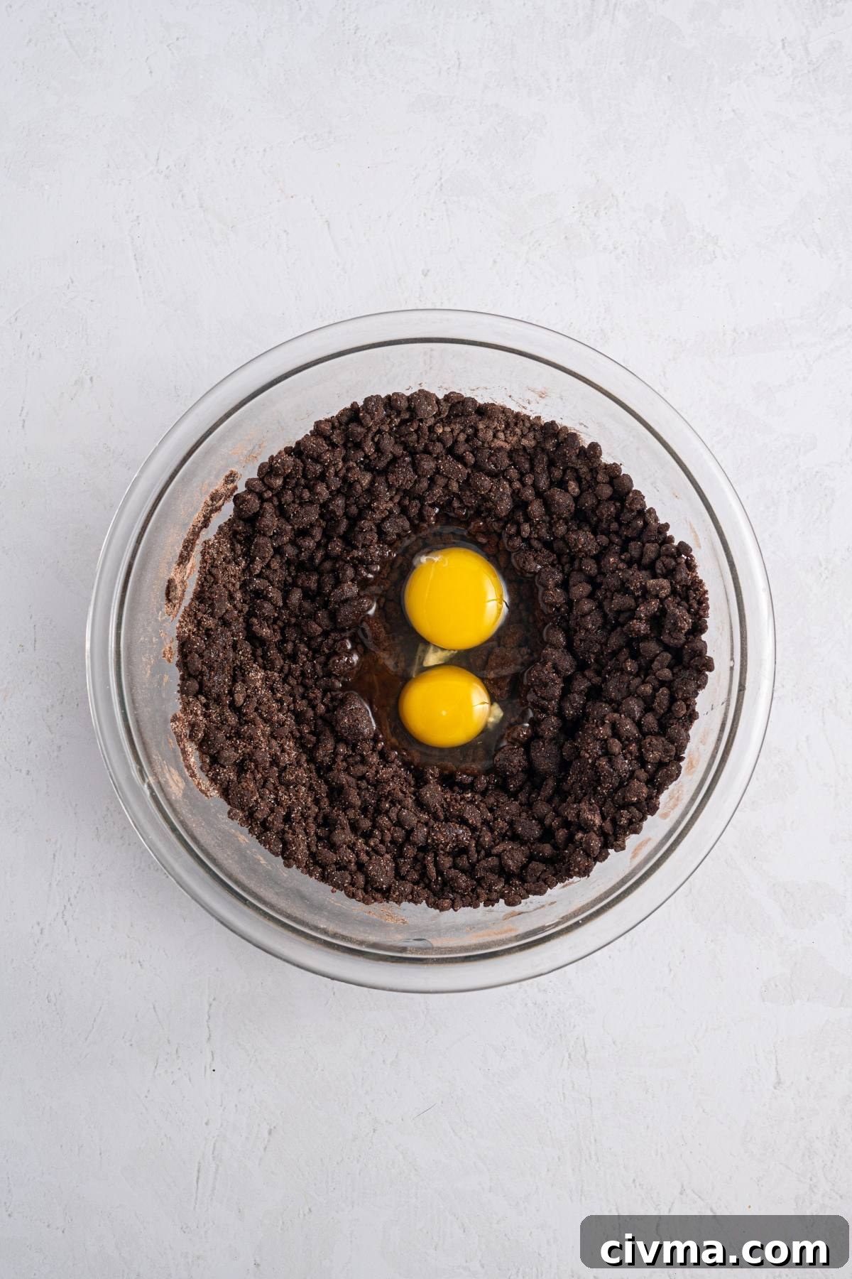A glass bowl with chocolate crumb mixture and two raw eggs in the center, on a white surface.