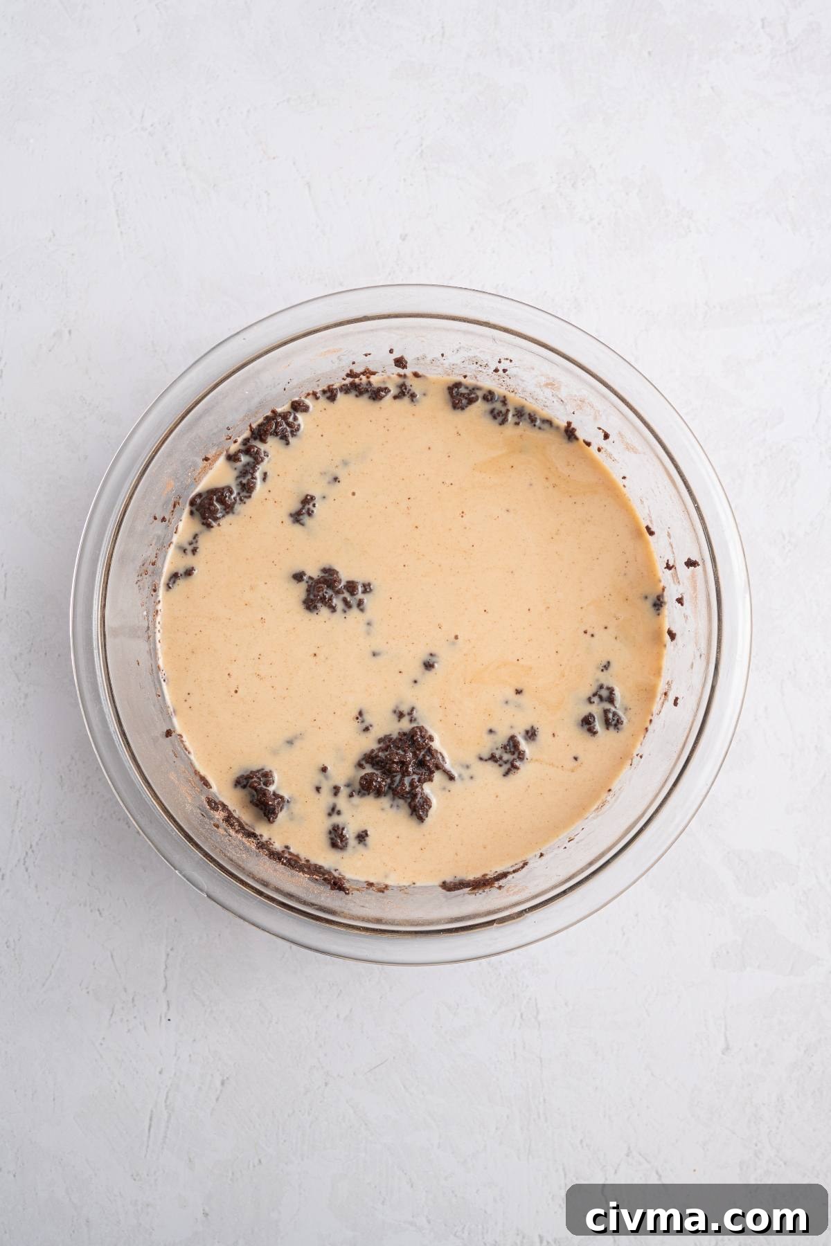 A glass bowl with a creamy batter and chunks of chocolate mixture on a light gray surface.