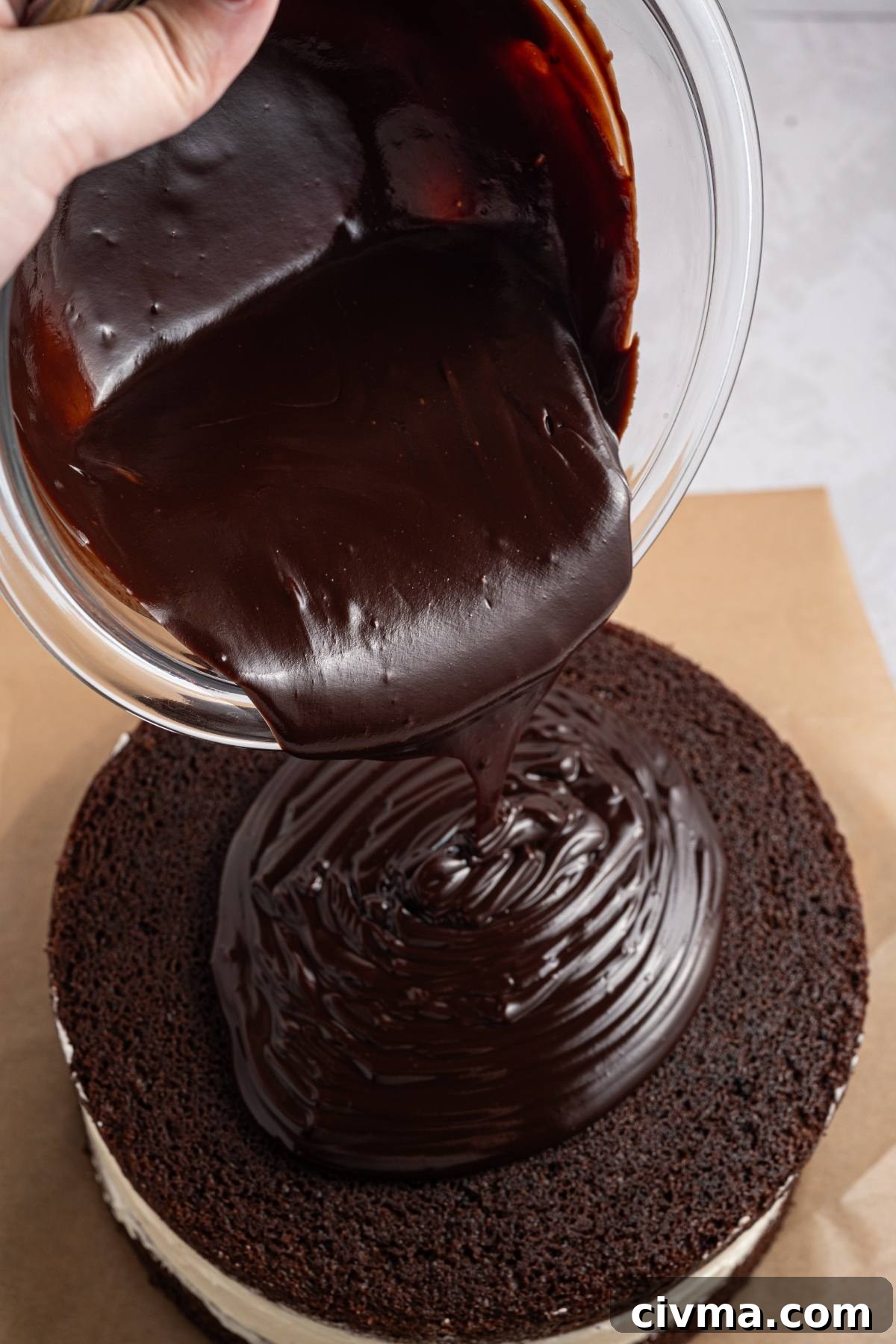 A bowl of chocolate ganache being poured onto a layered chocolate cake.