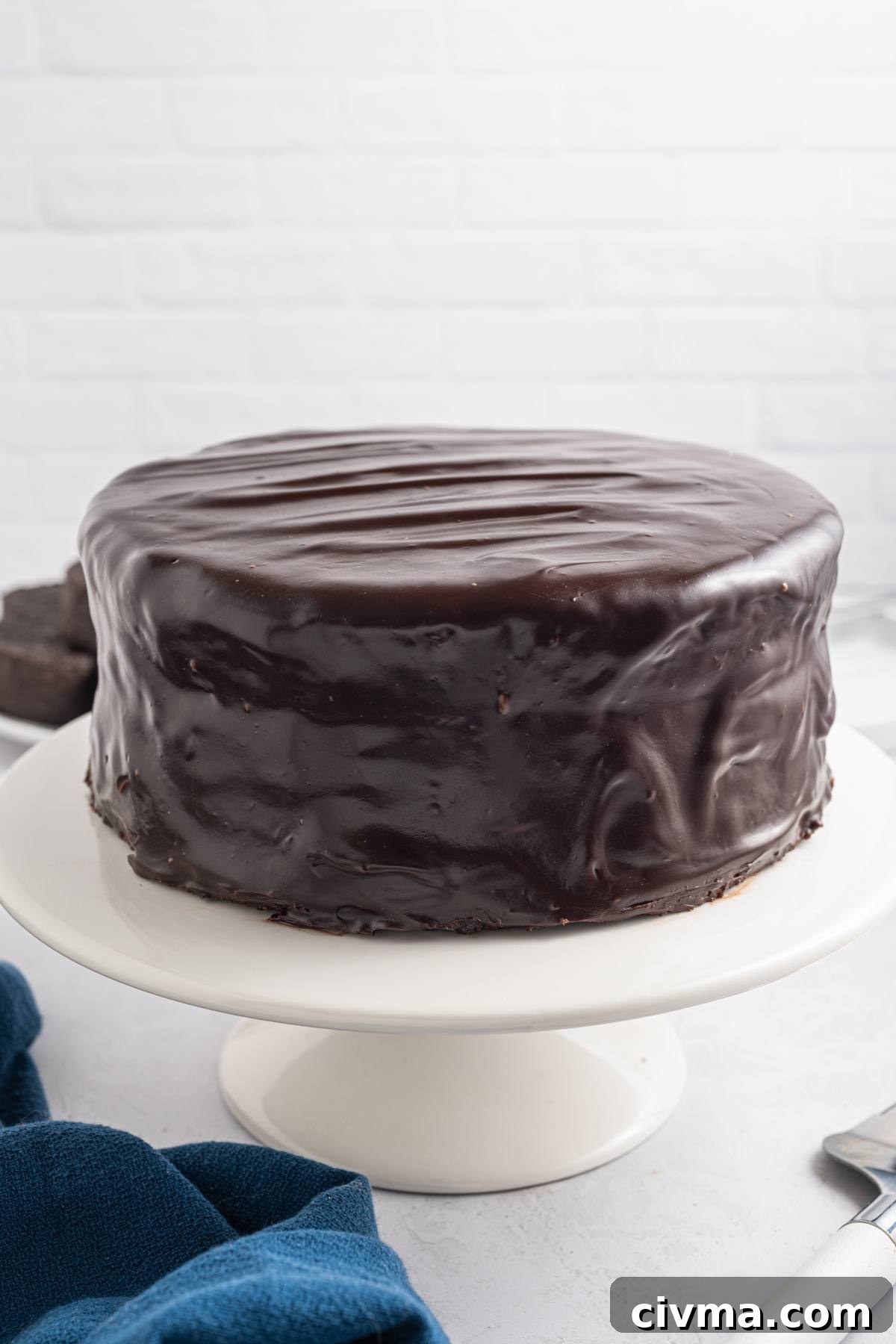 A round chocolate cake covered in glossy chocolate ganache on a white cake stand.