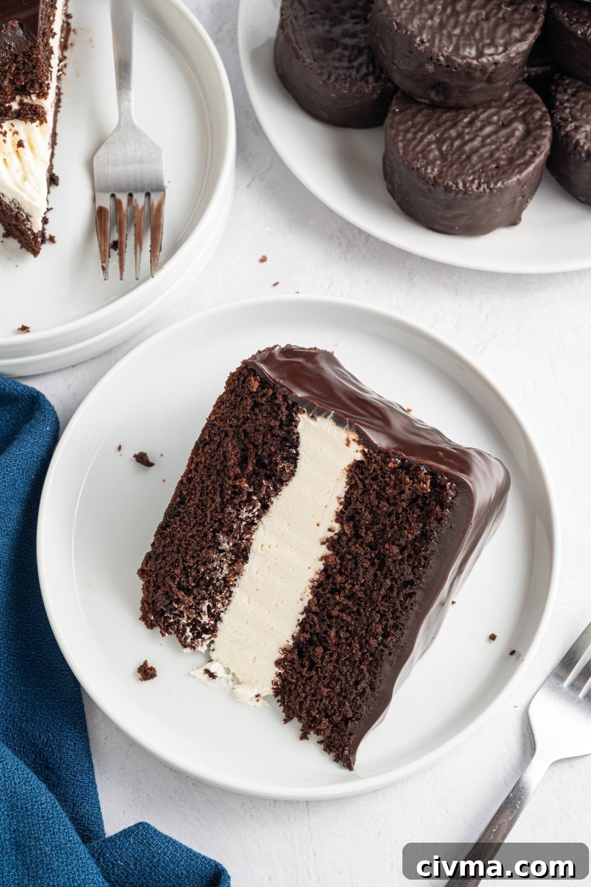 A slice of chocolate cake with cream filling on a white plate, next to chocolate-covered cookies.