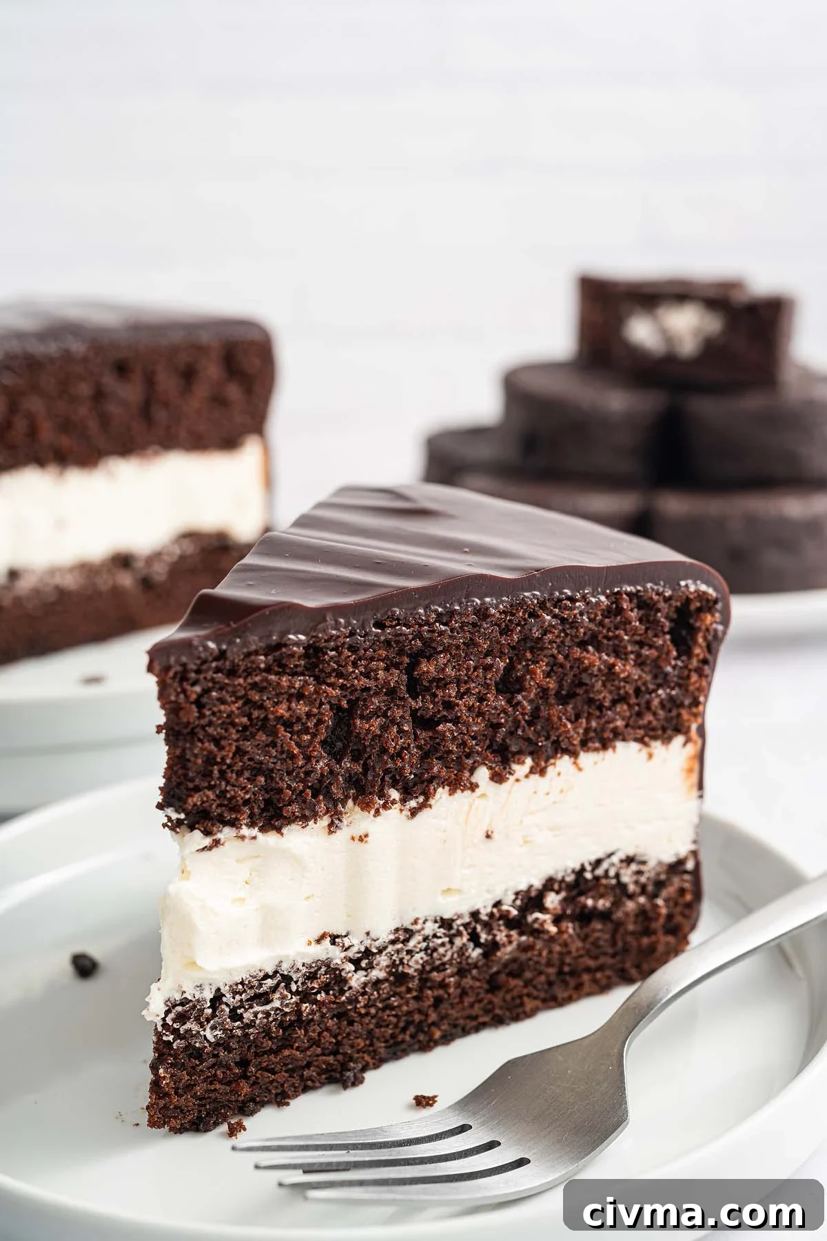 A slice of Ding Dong Cake with white cream filling, topped with rich chocolate glaze, sits on a white plate beside a fork.