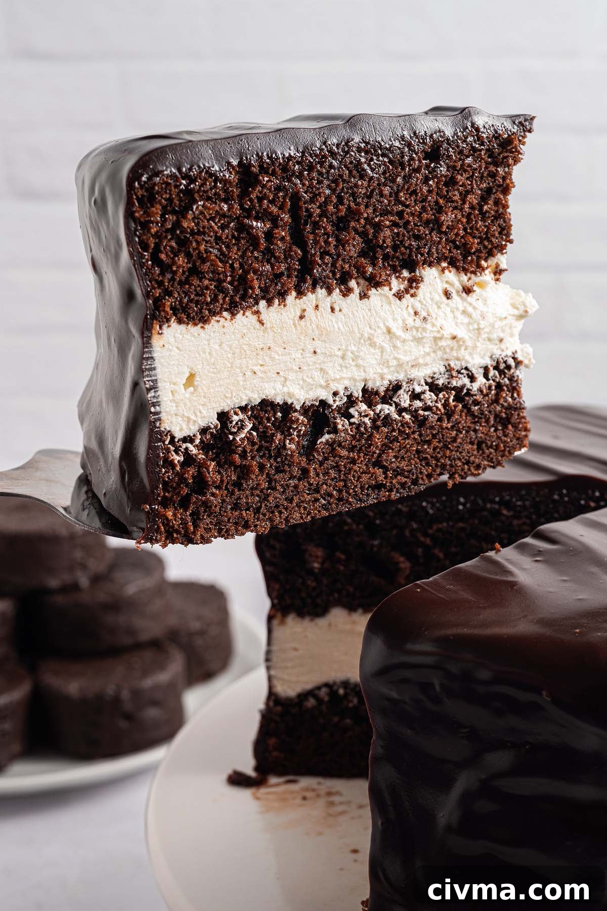 A slice of Ding Dong Cake with white cream filling and glossy chocolate frosting is being lifted from the cake.