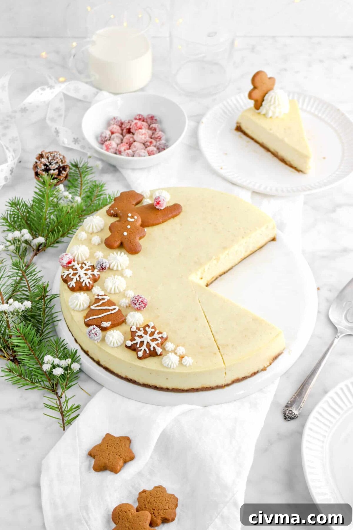 eggnog cheesecake with two slices removed, a slice behind on white plate, with pine branch, pinecone, bowl of cranberries, and mug of eggnog behind