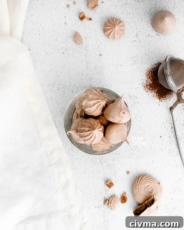 overhead shot of dark chocolate meringue cookies in a round glass cup on a white backdrop surrounded by a white towel, more chocolate meringue cookies, and a tea ball filled with cocoa powder