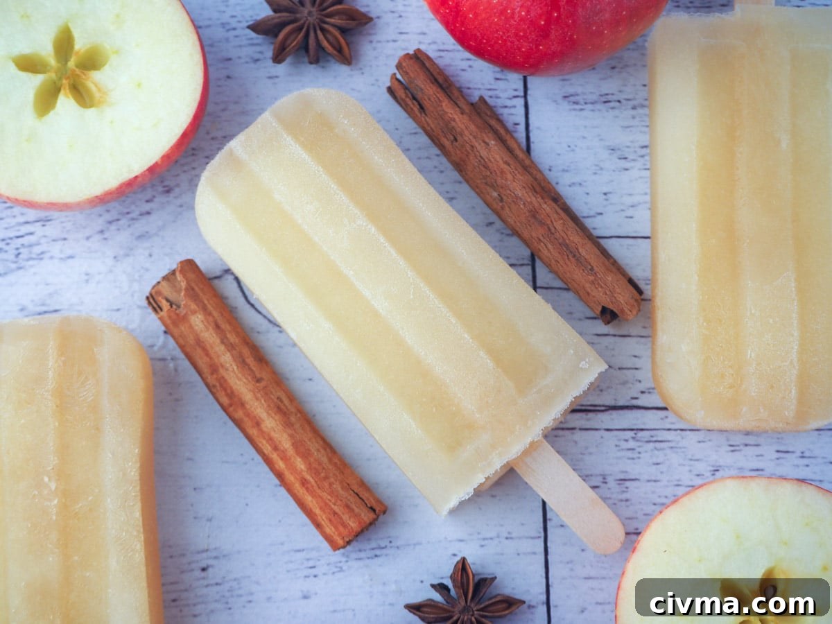 Stack of three mulled apple cider popsicles in front of two glass mugs of mulled apple cider, with apple stars and star anise in the foreground, on a chopping board.