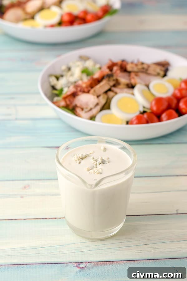 Dressing with blue cheese crumbled on top in a clear glass pitcher with two salads in the background