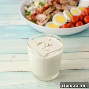 Dressing with blue cheese crumbled on top in a clear glass pitcher with two salads in the background - square