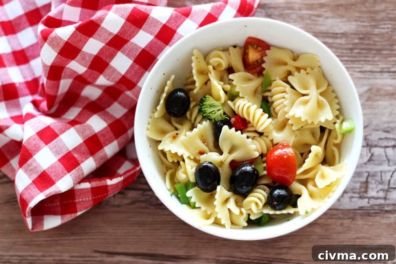 White bowl containing Italian Style Pasta Salad sitting on a brown table, red gingham napkin on table.