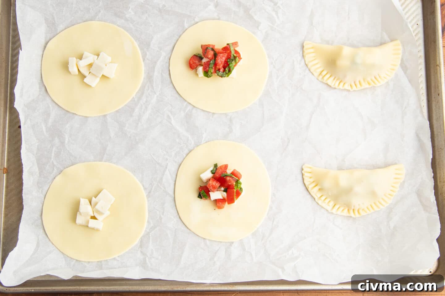 This is picture of a baking sheet lined with parchment paper and empanadas being assembled. 