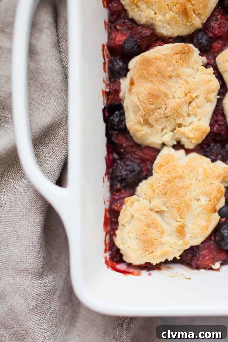 Homemade mixed berry cobbler in a white square baking dish with a tan napkin underneath.
