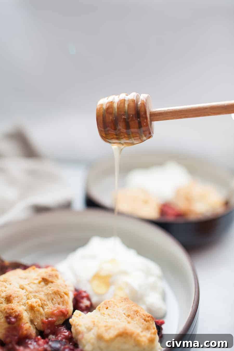 Honey pouring from a wooden honey dipper onto mixed berry cobbler with yogurt.