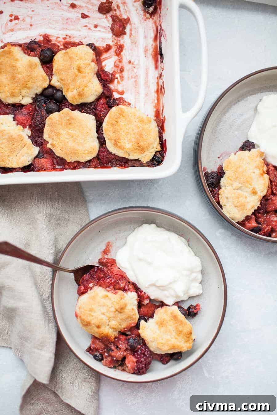 Overhead shot of two bowls of mixed berry cobbler with yogurt beside a white baking dish.