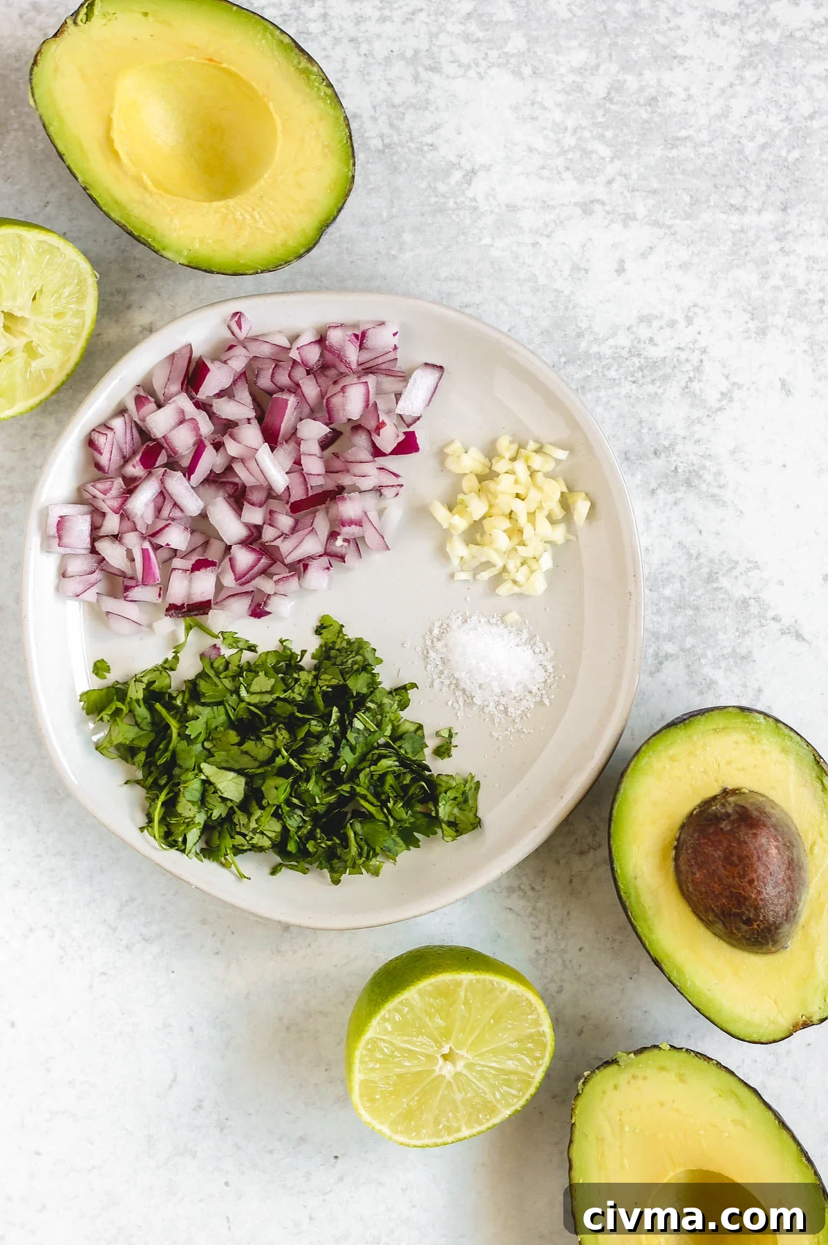 overhead view of cilantro, onions, garlic, and salt on a white plate with halved avocados and limes around the plate
