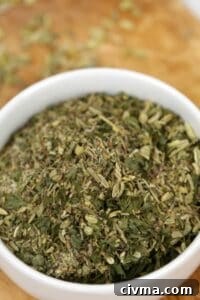 A white bowl filled with Porketta Seasoning, a mixture of dried green and brown herbs, sits on a wooden surface.