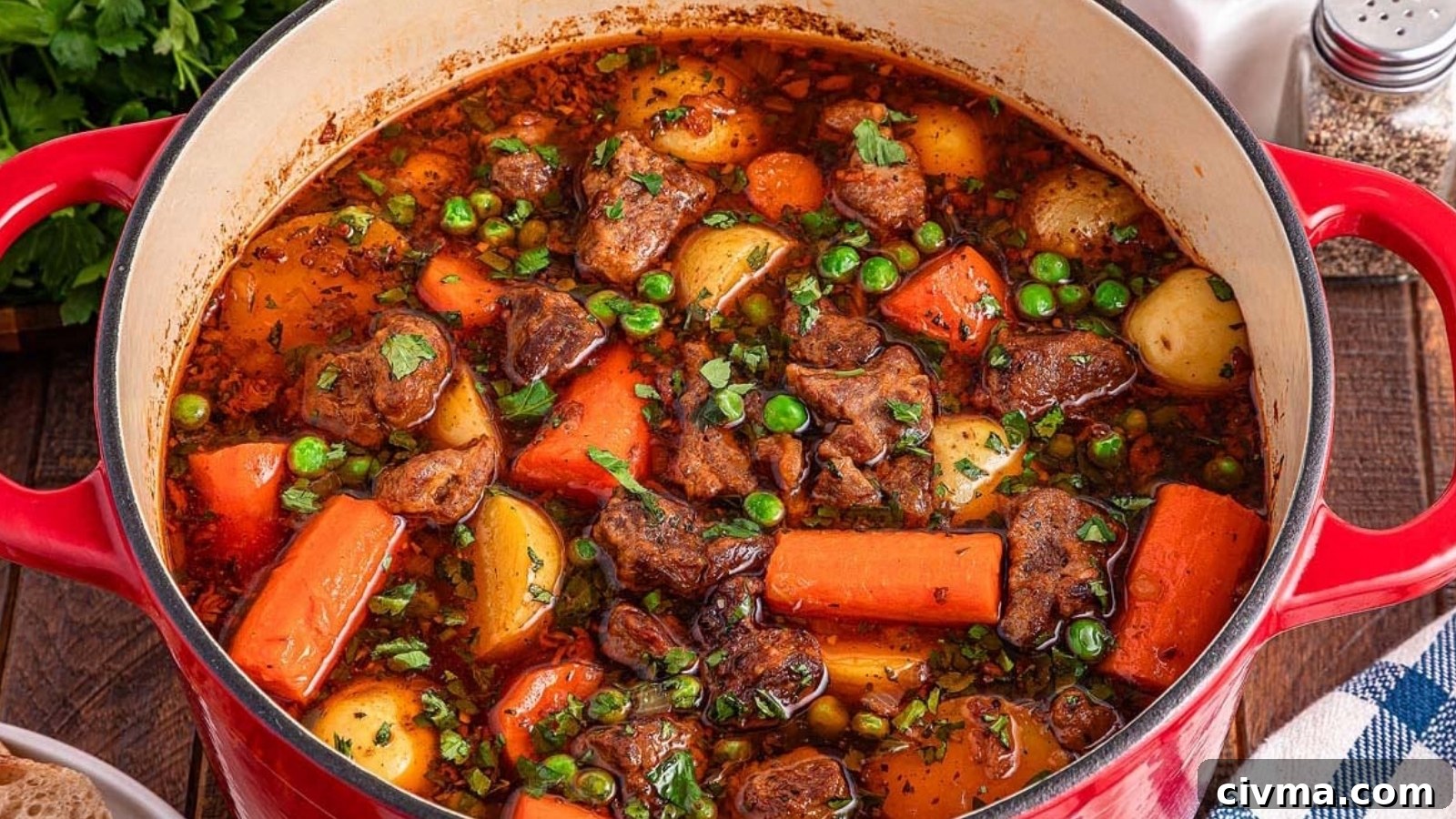 A red pot filled with beef stew containing chunks of beef, carrots, potatoes, peas, and herbs in a rich broth, sitting on a wooden table.