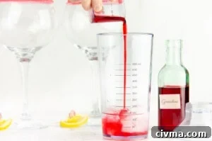 Poring grenadine into the gin in a cocktail shaker with two gin glasses, lemon slices and a green bottle of grenadine in the background