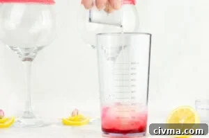 Pouring lemon juice into the gin and grenadine mixture in a clear plastic cocktail shaker with two sugar rimmed gin glasses and slices of lemon in the background