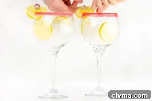Two sugar rim gin glasses with ice cubes and slices of lemons inside the glasses and placing lemon garnish with a rose petal onto the glass rim by hand