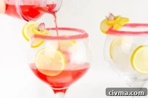 Pouring gin fizz into a large gin glass sugar rimmed garnished with lemon slice and rose petal with slices of lemon and ice cubes in the glass