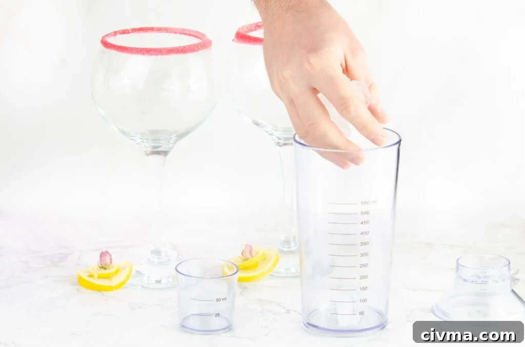 Placing cubes of ice by hand into a cocktail shaker with two sugar rimmed gin glasses and slices of lemon in the background