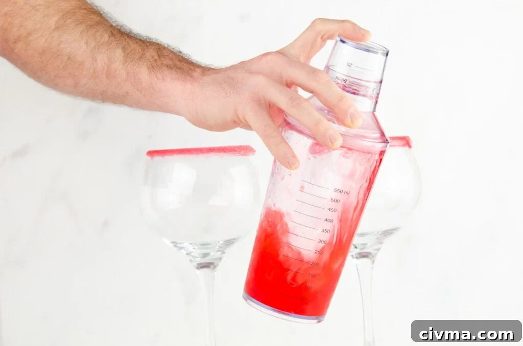 Shaking our pink gin fizz in a clear plastic cocktail shaker with two sugar rim gin glasses in the background