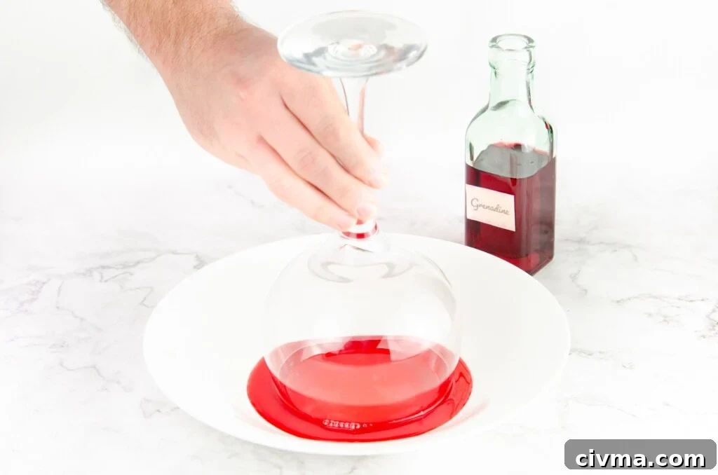 Dipping a gin glass rim into grenadine in a white bowl with a green bottle of grenadine in the background
