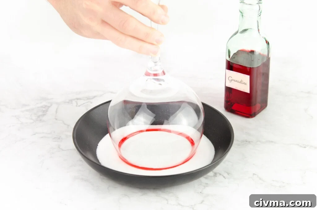 Dipping a gin glass coated in grenadine into caster sugar in a black bowl to make a sugar rim glass with a green bottle of grenadine in the background